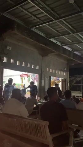 A group of people are gathered outdoors at night, possibly watching a screen that is not fully visible. There are several individuals standing and sitting. In the background, there appears to be a building with open doorways and shelves stocked with items, suggesting it might be a small shop or storefront. The ceiling structure above the gathering is visible, made of metal beams. The lighting is dim, indicating it is nighttime. Some individuals are looking towards the front, while others are facing away from the camera. The overall atmosphere seems to be social and potentially celebratory or engaged in a shared activity.