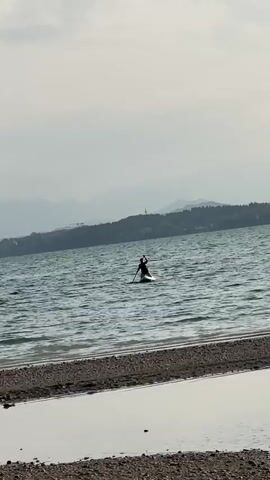A person is paddleboarding on a body of water. The person is standing on a paddleboard and holding a paddle. The person is facing away from the camera. The water is calm and there are waves. In the background, there are mountains and trees. The sky is cloudy. There is a beach in the foreground with pebbles and water.
