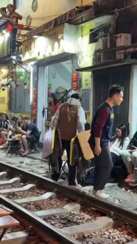 The video shows a busy street scene in Hanoi, Vietnam. People are sitting at tables on and next to a functioning railway track. A man is carrying two large, brown, rectangular objects that look like folded chairs. He carefully walks along the track, navigating between the tables and people. Other people are moving around, talking, and generally enjoying the street food and atmosphere. The area is brightly lit with decorative lights and the buildings are a mix of colors and styles. The camera slightly pans to follow the man carrying the chairs.