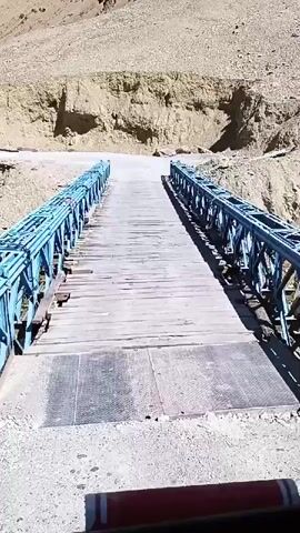 A video shows a truck driving across a metal and wooden bridge. The truck appears to be a large commercial vehicle, possibly an Indian or Pakistani truck, with a lot of decorations inside the cab. The driver, a man with a beard, is visible at the wheel. The bridge spans a rocky gorge, and the surrounding landscape is mountainous and arid. The video is shot from inside the truck's cab, looking forward. The bridge has blue metal trusses and a wooden deck. Some parts of the wooden deck appear worn or damaged. The video is short, about 4 seconds long, and the truck is moving across the bridge.