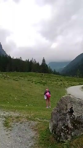 A low angle, wide shot captures a young girl standing in a grassy field near a dirt road. She is wearing a pink shirt, white shorts, and a backpack. The road curves into the distance, flanked by a steep, tree-covered mountainside. The sky is overcast, with a few patches of light filtering through the clouds. In the foreground, a large rock sits in the grass. The overall impression is one of a peaceful, natural landscape.
