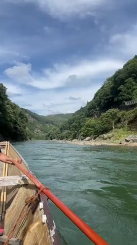 The video shows a first-person perspective from a wooden boat traveling down a wide, green river. The boat is equipped with a red-tipped wooden pole, presumably for steering or propelling the boat. A person's arm and shoulder, wearing a black t-shirt, are visible on the left side of the frame, holding the pole. In the background, lush green hills covered in trees rise steeply on either side of the river. The sky is blue with scattered white clouds, and the sun appears to be shining. The water is rippled, indicating movement. The overall scene suggests a serene and natural environment, likely a scenic river cruise.