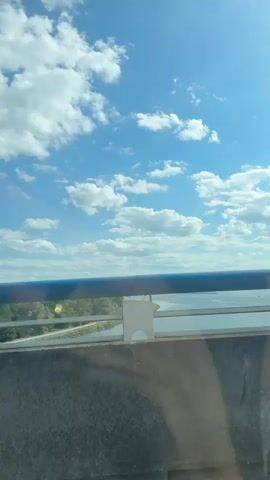 The video shows a first-person perspective from inside a vehicle, seemingly on a highway or bridge. The sky is a bright blue with scattered white clouds. The vehicle is moving and the camera captures the road ahead, the dashboard, and a rearview mirror. There are other vehicles on the road, including cars and a white van. The surroundings include a body of water, trees, and some industrial-looking structures with tall towers. The video also shows a blue railing along the side of the road. The text overlay indicates "Blijf 4 kilometer doorrijden," which translates to "Continue for 4 kilometers."
