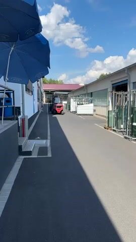 This image depicts an outdoor scene at what appears to be an industrial or manufacturing facility. The main subjects are a red forklift, several large glass panes stacked vertically on metal racks, and a blue umbrella in the foreground. The setting is an asphalted pathway or service road between buildings, with the buildings having industrial characteristics, including large windows and loading bays.

The forklift is positioned in the middle distance, facing towards the viewer. The glass panes are arranged along the right side of the pathway, suggesting they are either awaiting processing, storage, or transport. White rectangular objects, possibly crates or packaged materials, are also visible on the left side of the pathway, further down from the forklift.

The time of day is clearly daytime, indicated by the bright sunlight casting long shadows across the pathway. The weather appears to be fair and sunny, with a bright blue sky and scattered white clouds. The environment suggests a functional, perhaps busy, work site. There are no people visibly present in the frame.

The location cues, combined with the industrial nature of the site and the presence of specialized equipment and materials like glass, hint at an operation involved in glass manufacturing, processing, or distribution. The overall mood is neutral and observational, focusing on the industrial landscape under a pleasant sky. No legible text is visible in the image.