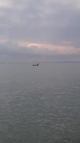 The video shows a body of water under a cloudy sky. The water is relatively calm, with small ripples visible on the surface. In the distance, there's a small silhouette that appears to be a boat or other watercraft. The sky is overcast with grey clouds, but there is a hint of light breaking through the clouds near the horizon, creating a pinkish glow.