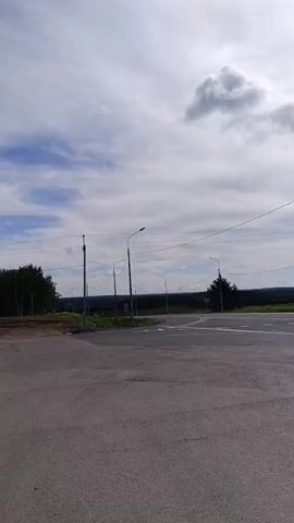 A black car makes a U-turn at an intersection in a rural area.  The intersection appears lightly traveled. There are street lights and road signs. The weather is cloudy with a hint of sun. The surrounding landscape is mostly green fields with some distant trees. There are power lines overhead.