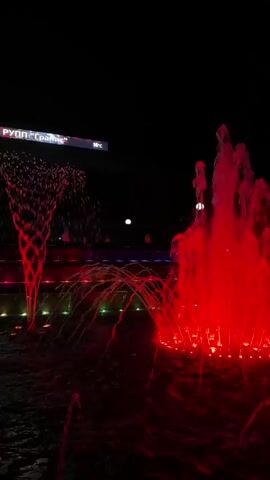 A nighttime video shows a fountain with red lights. The fountain has multiple streams of water that are illuminated by red lights. In the background, there is a sign that says "Грант" and some blurry lights. The water is splashing and creating a vibrant display.