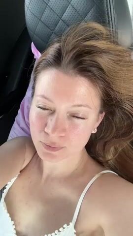 The video shows a woman in a car. She is wearing a white camisole and has her hair down. Her feet are propped up on the dashboard, and she is wearing white socks. She smiles at the camera and raises her hand. The car interior is visible, including the airbag warning label on the sun visor and the infotainment screen. The exterior view shows a blurred landscape of greenery and a dirt road.