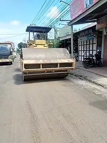 A road roller is compacting asphalt on a street. Several vehicles, including trucks and cars, are on the road. Buildings and power lines are visible on the sides of the street. The sky is clear and blue. The video is taken from a moving vehicle, likely a motorcycle, as evidenced by the bumpy camera movement. The video appears to be taken in a semi-urban or rural area during the daytime.