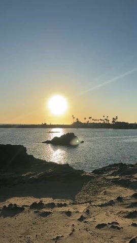 The scene captures a sunset view from a sandy beach, overlooking a calm body of water with a rock formation in the middle. The sun, large and bright, dominates the sky, casting a golden glare on the water's surface. In the distance, a coastline dotted with palm trees and buildings is visible. The foreground consists of sandy terrain with patches of darker rocky outcrops. The sky is clear, transitioning from a lighter blue near the horizon to a deeper blue overhead. There is very little movement, and the overall impression is one of tranquility and natural beauty.
