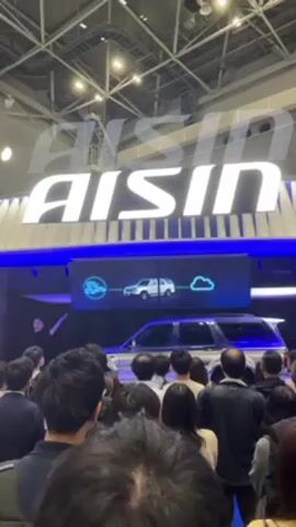 A crowd of people stand facing the camera at what appears to be a trade show or convention. Overhead, the AISIN logo is prominently displayed in bright white lettering. Below the logo, a wide screen shows a white SUV connected by lines to a cloud icon and a steering wheel icon. A real silver SUV sits below the screen with a blue glow emanating from beneath it. The lighting is bright overhead and appears to be indoor lighting. Most of the people in the crowd have their backs to the camera and are observing the display. The overall impression is that the video shows a demonstration of automotive technology being presented at a trade show by the company AISIN.