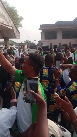A crowd of people is celebrating outdoors, most wearing green shirts with “CYON WARRI DIOCESE” printed on the back. Some people are recording with their phones held high above the crowd. A woman in the foreground is smiling broadly while recording. Buildings and cars are visible in the background.