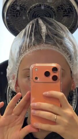 A woman wearing a clear plastic cap and a black salon cape sits in a salon chair, taking a selfie with her orange phone. Above her is a black and silver hair dryer with multiple fans. The shot is taken in a salon, reflected in a mirror. A window and some salon decor are visible in the background.