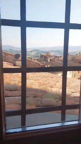 A medium shot captures a view through a window with a dark metal grille. The view looks out over a village or town, with a prominent expanse of terracotta-tiled roofs in the foreground. Beyond these roofs, more buildings with similar tiled roofs are visible, interspersed with some stone structures. In the distance, rolling hills under a clear blue sky suggest a rural or semi-rural setting. The light suggests it is daytime, likely late morning or early afternoon, with bright sunshine casting shadows and illuminating the landscape. The scene evokes a sense of peacefulness and timelessness, characteristic of many historic Italian towns. There is no visible text.