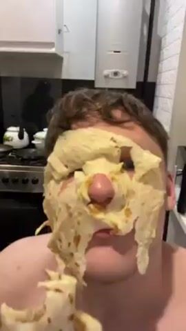A bare-chested man with brown hair has a tortilla-like substance stuck to his face, covering most of his head. A child's voice is heard offscreen. The man's mouth is slightly open.  A kitchen setting is visible with a stove, kettle, and water heater.  The man is holding the material in his hand.