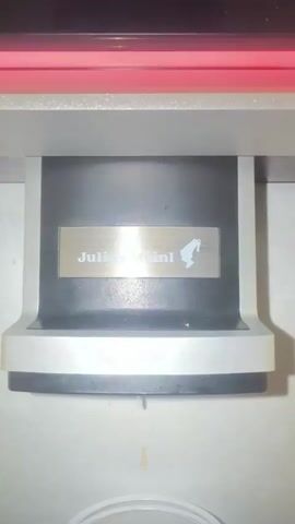 The video shows a close-up, top-down view of a coffee machine dispensing two streams of liquid into clear cups below. The machine has a black central unit with a silver plate bearing the logo "Julius Meinl" and an emblem of a horse. Above the dispensing mechanism, there is a red light. The machine is mounted within a grey housing, and the background appears to be a light-colored, textured surface. The dispensing begins at 00:03, with liquid flowing steadily from two spouts.