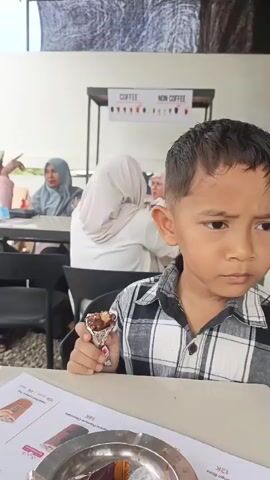 A young boy in a plaid shirt holds a snack, looking off to the side. Behind him, a few women are seated at tables, some wearing hijabs. In the background, a sign displays "COFFEE" and "NON COFFEE." The scene appears to be an outdoor seating area, possibly at a cafe or food stall. Further in the background, a gas station with red and white accents is visible, with vehicles parked around it. A girl is seen at a table in the mid-ground, and a woman is looking at her phone at a table in the background.