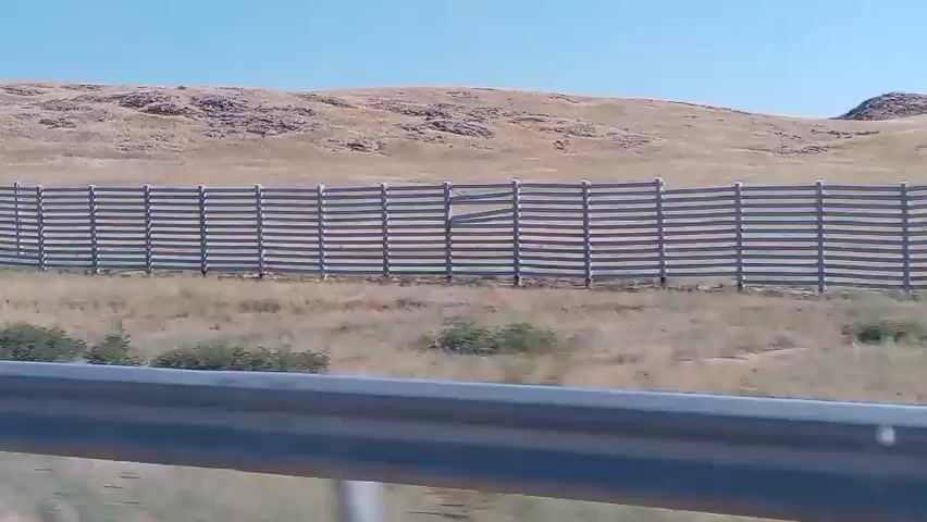 The video captures a wide shot of a desolate, dry landscape under a clear blue sky. In the foreground, a silver guardrail runs horizontally, slightly out of focus. Beyond the guardrail, a field of dry, brown grass stretches into the distance. A wooden fence, constructed from horizontal planks supported by vertical posts, dominates the mid-ground, curving gently to the right. To the left of the wooden fence, a series of individual wooden posts are spaced evenly, extending towards the horizon. In the background, rolling hills, covered in sparse, dry vegetation, rise against the sky. The overall impression is of a vast, open, and somewhat barren environment. The camera appears to be moving, suggesting the footage was recorded from a vehicle.