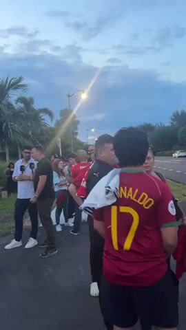 The video shows a group of people gathered outside, likely near a street or pathway. The lighting suggests it is either dusk or dawn, with a cloudy sky. Most of the individuals are wearing red shirts, many of which appear to be soccer jerseys with the name "CRISTIANO" visible on the back of one. One person in the foreground has "RONALDO 17" on their back. Several individuals are talking and interacting with each other. There are palm trees visible in the background, and what appears to be a street lamp. The overall atmosphere seems to be one of anticipation or post-event gathering related to a sporting event.