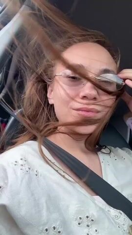A woman with long, brown hair is in the passenger seat of a car. Her hair is blowing in her face. She is wearing glasses and a white shirt with eyelet details. The seatbelt is visible. The background is blurred.