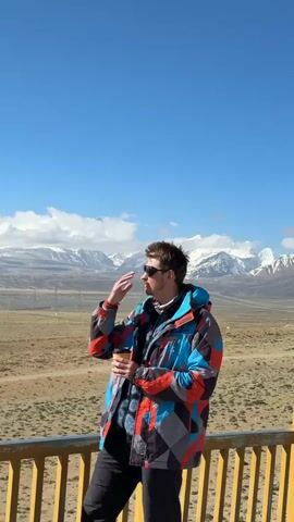 In a bright outdoor scene set in Gyirong County, China, a man stands on what appears to be a wooden balcony or viewing platform. He is seen wearing a vibrant winter jacket featuring a geometric pattern in shades of blue, red, grey, and black, paired with black pants and sunglasses.

At the beginning of the video (0:00), he is seen shielding his eyes with one hand, looking out towards the expansive landscape, while holding a yellow coffee cup in the other. He then lowers his hand and, from 0:01, brings the cup to his lips, taking a sip from the drink. The text "COFFEE" is visible on the cup, and the brand "PROTEST" can be subtly seen on his jacket.

The background reveals a breathtaking panoramic view under a clear blue sky dotted with scattered white clouds. Majestic snow-capped mountains stretch across the horizon, contrasting with the vast, dry, and somewhat barren high-altitude terrain in the middle ground, which features sparse, low-lying vegetation. Some distant structures, possibly power lines, are also visible. The bright daylight suggests a clear morning or early afternoon. The man appears relaxed, seemingly enjoying both his beverage and the impressive mountainous scenery.
