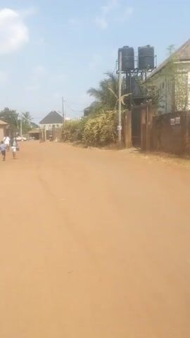 The video shows a dirt road in a residential area. The road is a reddish-brown color and appears to be unpaved. On the right side of the road, there's a weathered wall with a metal gate, behind which are two black water tanks elevated on a metal structure. Lush green foliage grows along the wall. On the left side of the road, a few people are walking in the distance. Some buildings are visible in the background, including a house with a dark roof and a lighter-colored building further down the road. Power lines are strung between poles in the distance. The sky is blue with scattered white clouds.