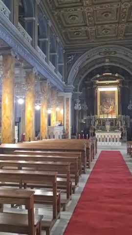 The video shows the interior of a church. In the foreground, there are rows of wooden pews on the left and right sides of a red carpet runner that leads towards the altar. The altar is at the far end of the church and features a large painting above it. The church has high ceilings with intricate detailing and ornate columns lining the sides of the nave. Chandeliers hang from the ceiling. The overall lighting suggests it is daytime, but the interior is dimly lit. There are no people clearly visible in the shot.
