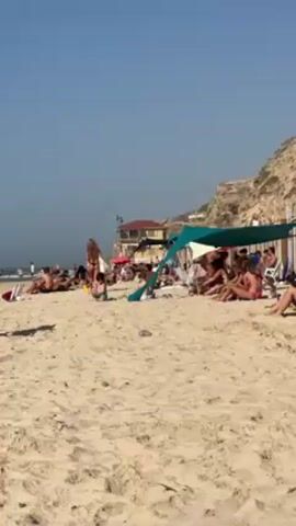 The video shows a crowded beach on a sunny day. People are sunbathing and relaxing on the sand, some under a turquoise-colored canopy. A child is playing near the water. In the background, there's a building and a cliff. The sand is light-colored, and the sky is clear blue.