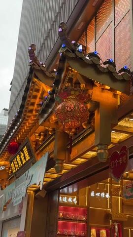 The video shows a close-up of a Chinese lantern hanging from a golden, ornate roof structure. The lantern is red and gold with intricate details and several levels. Characters are visible on the lantern. Small, colorful lights decorate the roof structure, which appears to be part of the facade of a building. A sign with Chinese characters is also visible. The background shows a modern building.