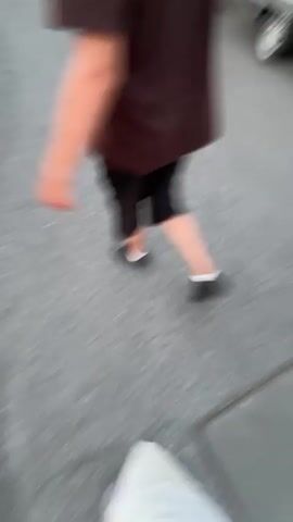 The media file captures a candid moment on a paved street in Oelde, Germany. The primary subjects are two individuals walking, their forms slightly blurred due to motion. One person, seen from the waist down, wears a dark brown t-shirt and black shorts, with dark shoes and white socks. The other individual, also captured from the legs down, is wearing shorts with a distinctive checkered pattern and athletic shoes. Their legs also show visible tattoos.

In the background, a white trailer with visible wheels is parked alongside the street. A dark-colored Audi is also parked nearby. The scene appears to be outdoors, with hints of residential buildings and houses visible in the distance, suggesting a suburban or semi-urban setting. The lighting suggests it is daytime, possibly late afternoon or early evening, given the soft light and the absence of harsh shadows. The weather appears to be fair.

The activity is simply walking along the street. There are no overt signs of distress or excitement; the mood seems relaxed and casual. The individuals appear to be interacting or walking together. The visual information is limited to the lower bodies and immediate surroundings of the walkers, with the background providing context about the location and environment. No visible text is present in the media.