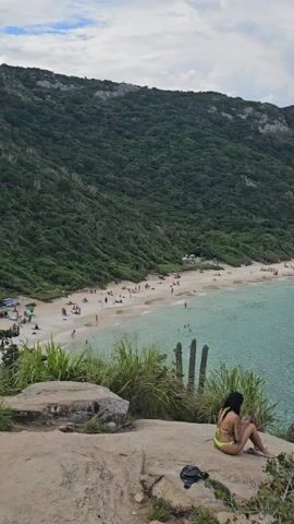 The video shows an overview of a beach and bay.  In the foreground, a woman in a yellow bikini sits on a rock, using a phone.  The shot pans to the right, revealing a sandy beach with people scattered along it, and then a large bay with turquoise water, surrounded by hills covered in green forest.  A tour boat is visible in the bay, sailing from left to right. Cactuses grow on the rocky outcrop where the woman is sitting. The sky is overcast.
