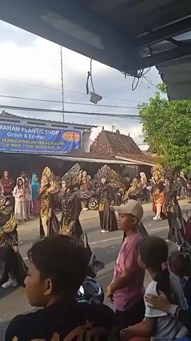 People are lined up on a street watching a parade.  The parade features performers in elaborate, dark-colored costumes with gold accents. Some of the costumes have large headpieces or wings. Spectators of all ages are visible, including children and adults, some of whom are taking photos or videos with their phones. The street is lined with buildings, and there are overhead power lines. There is a sign for "GRAHAM PLASTIC SHOP" visible. The sky is overcast. The camera is positioned under an overhang, possibly a shop awning.
