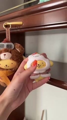 A person is holding a small, yellow, plush keychain toy in their hand. The toy has a white and brown hooded jacket on it. They are pressing on the toy, which makes a popping sound. Another keychain toy with the character Pompompurin dressed as a bear can be seen in the background hanging from a hook on a dark wooden cabinet. The person has reddish-orange nail polish on their nails.