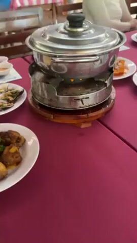 The video shows a table set with a burgundy tablecloth. There are several plates of food, including one with chicken pieces garnished with parsley, and another with clams or oysters topped with chopped nuts and herbs. In the center of the table is a stainless steel pot sitting on a heating device with a lit flame underneath. There are other dishes and condiments scattered around the table. Wooden chairs surround the table, and people are visible in the background, suggesting a restaurant or dining setting.