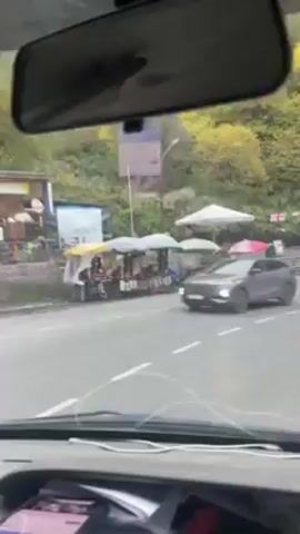 The video shows a car driving on a winding road in a mountainous area. The road is paved and has white lines marking the lanes. There are trees and foliage on either side of the road. Other cars are visible driving in the same direction. There are market-like stands along the road. The view is from inside the car, through the windshield and the rear-view mirror is visible. A white audio cable is lying on the dashboard. There is music playing that seems to be religious in nature.