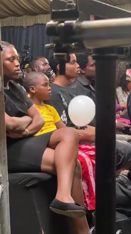 A group of people are sitting in an indoor venue. In the foreground, a young person in a yellow shirt sits with their arms crossed, holding a white balloon. To their left, a woman in a black dress is also seated. Behind them, several other individuals are visible, some looking forward. The environment appears to be a seated audience area, possibly for an event or performance, with a fabric ceiling visible above.