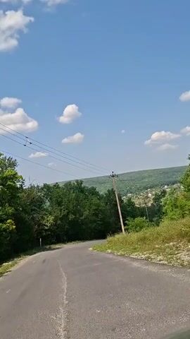 The video shows a rural road with a slightly curved path. On the left side of the road, there are many trees and dense foliage. On the right side, there is a grassy slope and a utility pole with wires extending across the road. The sky is blue with scattered white clouds. In the background, there are more trees and a distant view of houses on a hill. The camera appears to be moving forward along the road.