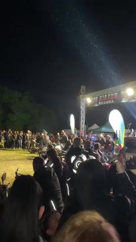 At night, a crowd has gathered in an open area, likely a festival. The ground is dirt. A stage with bright lights and signage is visible in the background. The sign reads “BULGARIA” at the top. People are standing in front of several motorcycles, some wearing jackets with reflective designs. Many in the crowd are taking pictures with their phones. Trees are on the edge of the area. It appears to be raining.