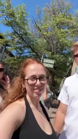 The video shows three young adults walking on a sidewalk, seemingly taking a selfie video with a phone held by a red-haired woman wearing glasses and a black halter top. Another young woman with blonde hair, sunglasses, and a white tank top walks beside her. A blonde man in a white t-shirt walks alongside them, momentarily appearing in the frame. The backdrop consists of trees, buildings, and street signs, suggesting an urban or town environment on a sunny day.