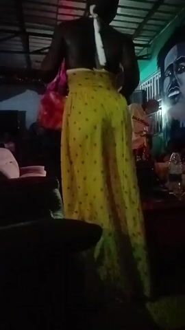 The video shows the back of a person in a club or similar venue. They are wearing a black top and a long, colorful skirt with dark heart patterns. The skirt changes color intermittently, flashing yellow, green, and orange. In the background, a person with a pink hat and red patterned shirt is partially visible.  A mural of a man’s face is also present on the wall.  There are a few indistinct people in the dark background and some tables with objects on them.  The lighting is dim.