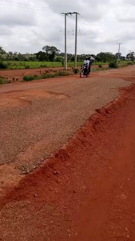 A video captures a dirt road scene with a motorcycle carrying three people, moving from left to right. The road is a mix of gravel and red dirt, with some visible potholes. The background features lush green vegetation, trees, and a few scattered houses with thatched roofs. Utility poles with wires are also present. The sky is overcast with gray clouds, suggesting a cloudy or potentially rainy day. The lighting appears to be natural daylight, possibly late morning or early afternoon. The overall atmosphere is rural and suggests a moment of transportation or travel in a Nigerian setting, specifically Sabon Sarki. No visible text is present in the video. The people on the motorcycle appear to be casually dressed.