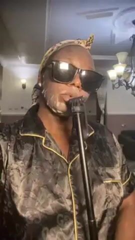 A person wearing black sunglasses, a patterned pajama shirt with gold trim, and a headscarf is holding a microphone. A white substance appears to be applied to their face, resembling a face mask. The background shows a room with a chandelier and what appears to be a wall sconce. The person says, "I'm going to sing for you guys a song. Stay tuned."