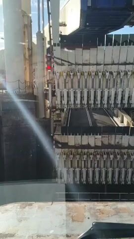 This video captures a close-up view of a ferry loading ramp, likely at the Dover, United Kingdom port. The focal point is a series of heavy-duty hydraulic mechanisms that appear to be part of the system for raising and lowering the ramp. These mechanisms are arranged in two horizontal rows, each with multiple cylindrical components and connecting rods. The ramp itself, a dark, textured surface, is visible between the rows of machinery.

The scene is set against a backdrop of industrial structures, including large white panels and beams, suggesting a robust, operational port environment. Overhead, a blue and white structure, possibly part of the ferry or the port's infrastructure, is visible. Sunlight is strongly present, indicated by a lens flare across the left side of the frame, suggesting it is daytime. The sky appears clear.

The activity captured is the stationary state of the loading ramp's mechanical components. There are no people visible, and the machinery appears to be at rest. The overall impression is one of powerful, industrial equipment. There is no visible text in the frame. The lighting and the sheer scale of the machinery suggest a busy, functional port.