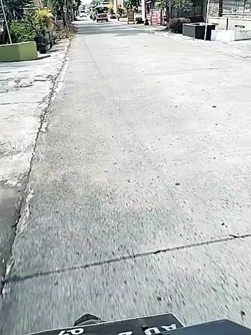 The video shows a grey asphalt road in a rural area. Trees and power lines line both sides of the road. A motorcycle with a driver wearing a white shirt drives towards the camera. The road is flanked by houses and signage. The weather appears sunny.