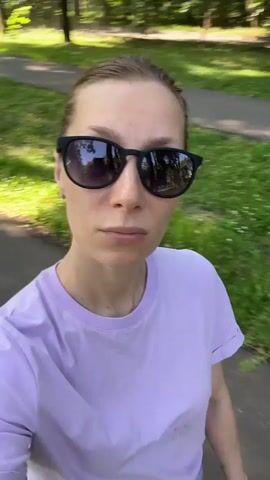A medium shot shows a woman from the chest up as she walks forward. She is wearing sunglasses and a purple t-shirt. The background is a park with trees and green grass.