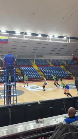 The video shows a volleyball game in progress inside a large, mostly empty stadium. Several players wearing green and blue uniforms are visible on the court. One player spikes the ball over the net, and her teammates celebrate after the point is scored. A referee stands on a ladder to observe the game. The stadium seating is primarily blue with some yellow sections. A Venezuelan flag is visible near the ceiling. An advertisement for SUR FILAS I-N can be seen on the wall of the stadium. Observers can be seen sitting near the camera in the foreground.