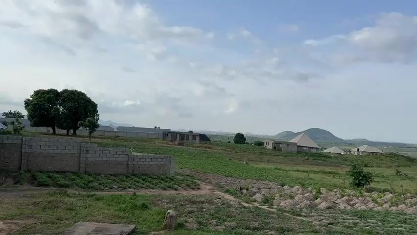 The video captures a panoramic view of a rural landscape under a partly cloudy sky. In the foreground, a low brick wall defines a construction site with incomplete buildings made of concrete blocks. Beyond the wall, rows of green crops stretch across the land, interspersed with patches of dry, tilled soil. A dirt path winds through the fields, leading towards the center of the frame. Several unfinished and completed houses are visible in the mid-ground, some with pitched roofs. The structures vary in their stage of construction, with some appearing as bare skeletons of concrete and others more finished with walls and roofs. In the distance, rolling green hills rise, some topped with rocky outcrops. A large, dense tree stands prominently in the middle ground, and a few other scattered trees dot the landscape. Further in the background, a town or city can be seen extending across the horizon, with more distant hills visible behind it. Power lines are also present in the distance, indicating infrastructure. The overall impression is one of development and settlement in a natural, hilly environment.