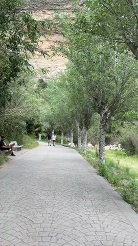 The video shows a paved pathway in a natural setting. A man sits on a bench on the left side of the path, seemingly occupied with something on his lap. Further down the path, two people walk together in the same direction. As the video continues, a man walks into the scene from the right, walking along the path towards the camera, carrying a bag in each hand and wearing a backpack. The path is lined with trees on both sides and there is a steep, rocky hill visible in the background. The environment appears to be a park or recreational area.
