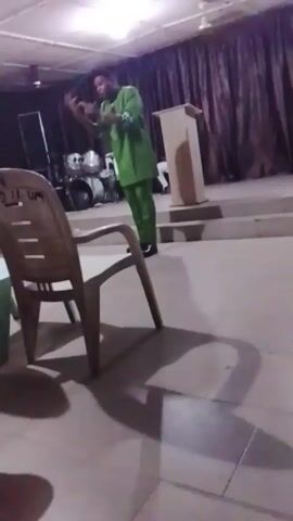 A man in a green suit is standing on a stage in front of a light wood-colored pulpit. He is speaking into a microphone and gesturing with his hands, at one point raising his right hand in the air. Behind him are drums and a dark curtain. In the foreground are two plastic chairs, the first one mostly beige with “18” and some illegible writing on the back. The room is brightly lit with fluorescent ceiling lights.