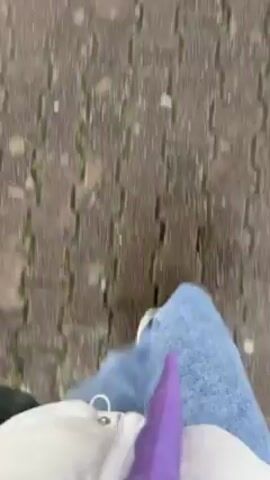 The video is taken from the perspective of a person walking on a wet brick sidewalk under an umbrella on a rainy day. The person is wearing light blue jeans and white sneakers. The sidewalk is lined with bare trees on one side and green grass and more trees on the other. Cars are parked along the road on the right. The umbrella is black with a gray checkerboard pattern on the inside. The sky is overcast and gray.